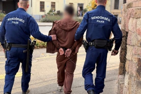 Két rendőr megbilincselt, kitakart arcú férfit kísér el