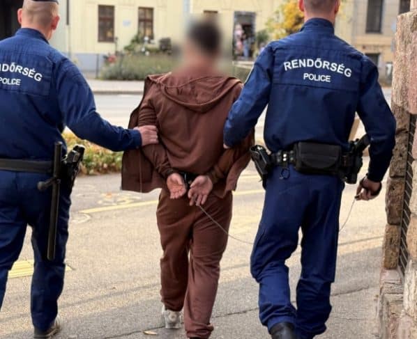 Két rendőr megbilincselt, kitakart arcú férfit kísér el