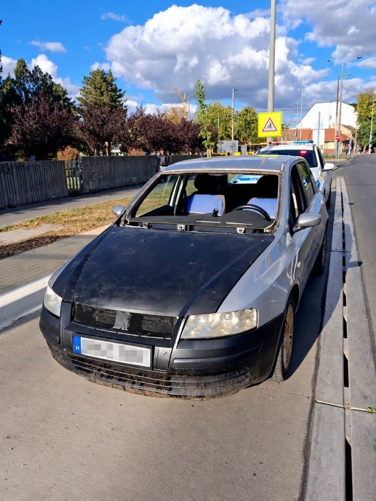 Szélvédő nélküli ezüst személyautó egy rendőrautó előtt Debrecenben, kitakart rendszámmal