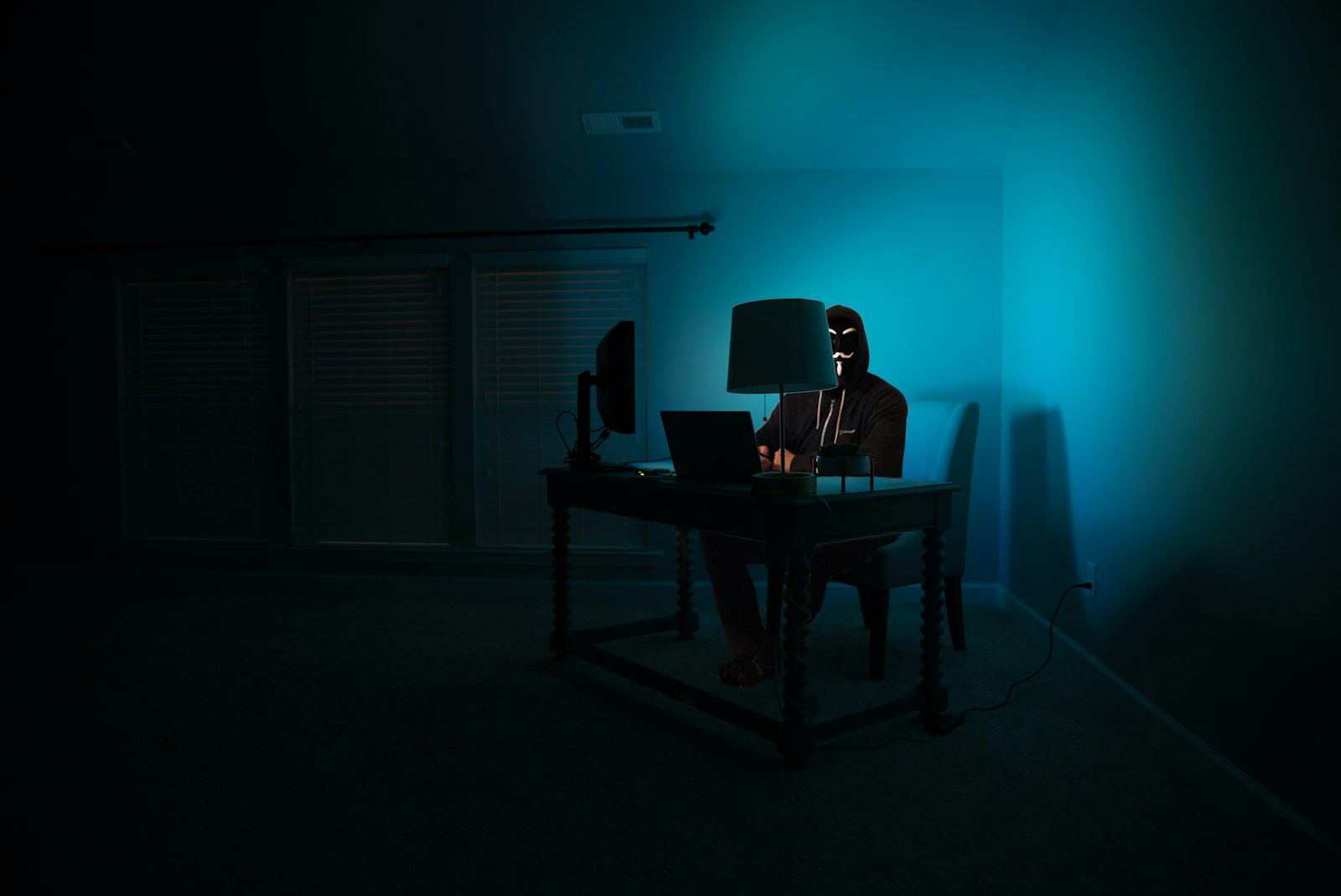 Kapucnis, maszkos alak laptop előtt sötét szobában – hackertámadás illusztráció